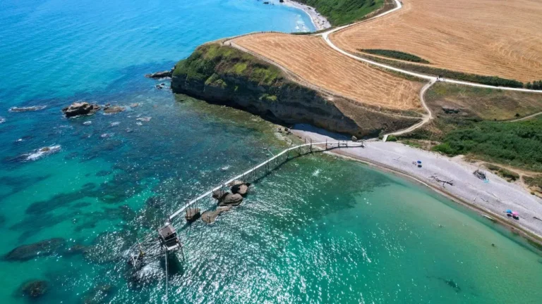 Veduta aerea di un trabocco su un mare cristallino turchese lungo la costa dell'Abruzzo, con sentieri panoramici e scogliere dorate.