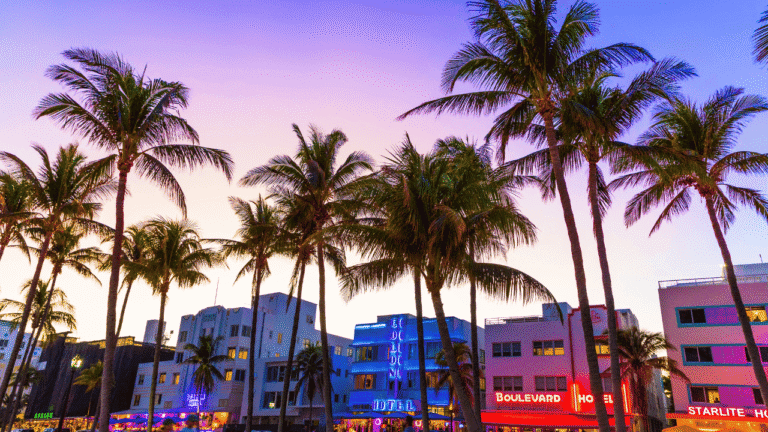 Veduta aerea di Miami Beach con l'ampia spiaggia dorata, le caratteristiche torrette colorate dei guardaspiaggia in stile Art Déco e l'oceano Atlantico che sfuma dal verde smeraldo al blu profondo.