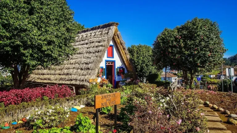 Case tradizionali di Santana a Madeira con i caratteristici tetti spioventi in paglia, pareti bianche e infissi rossi e blu, circondate da un giardino fiorito durante il tour di Explora nel Mondo.